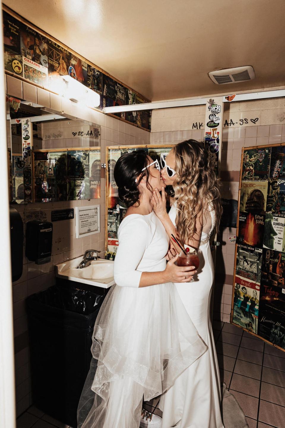 Two brides kiss in a bathroom wearing sunglasses and wedding dresses.
