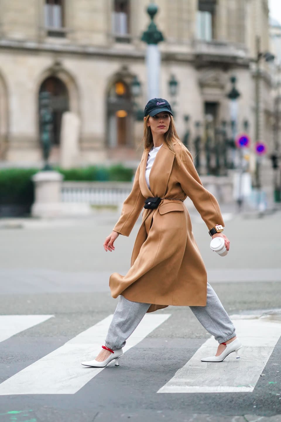 paris, france february 01 natalia verza aka mascarada wears a black cap from nike, a white t shirt, a beige pale brown wool long trench coat, gray sport jogger pants, a golden prada bracelet bag, a mini black quilted chanel belt bag, white square toe shoes with red features from prada, holds a prada hot drink coffee paper cup, on february 01, 2021 in paris, france photo by edward berthelotgetty images