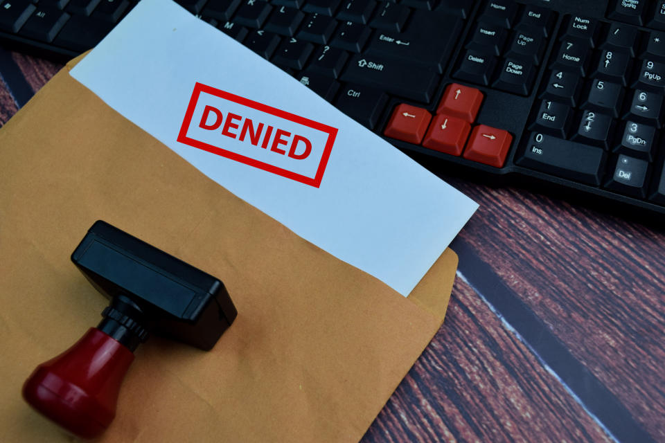 A stamped envelope with "DENIED" in red text is placed on a wooden surface next to a keyboard