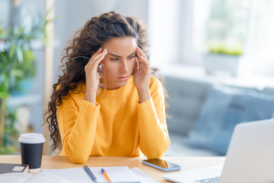 Young Latina woman in yellow rubbing her temples while holding her glasses trying to remember her password.