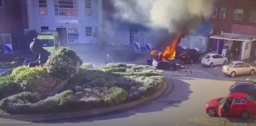 Screenshot of a video taken from a window looking down at a car on fire near a green roundabout. Cars can be seen parked around the roundabout near a sign that says reception.