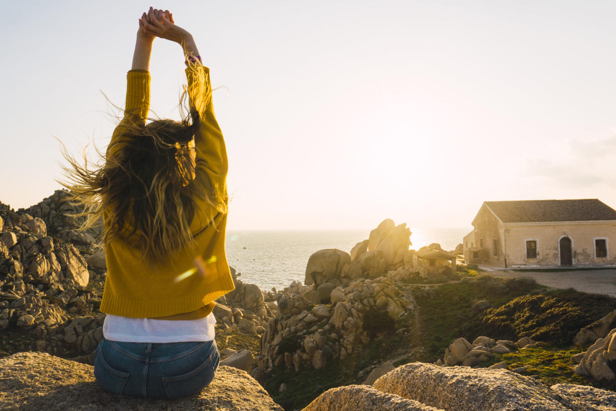 Italy, Sardinia, woman on a hiking trip sitting on rock at the coast raising her arms