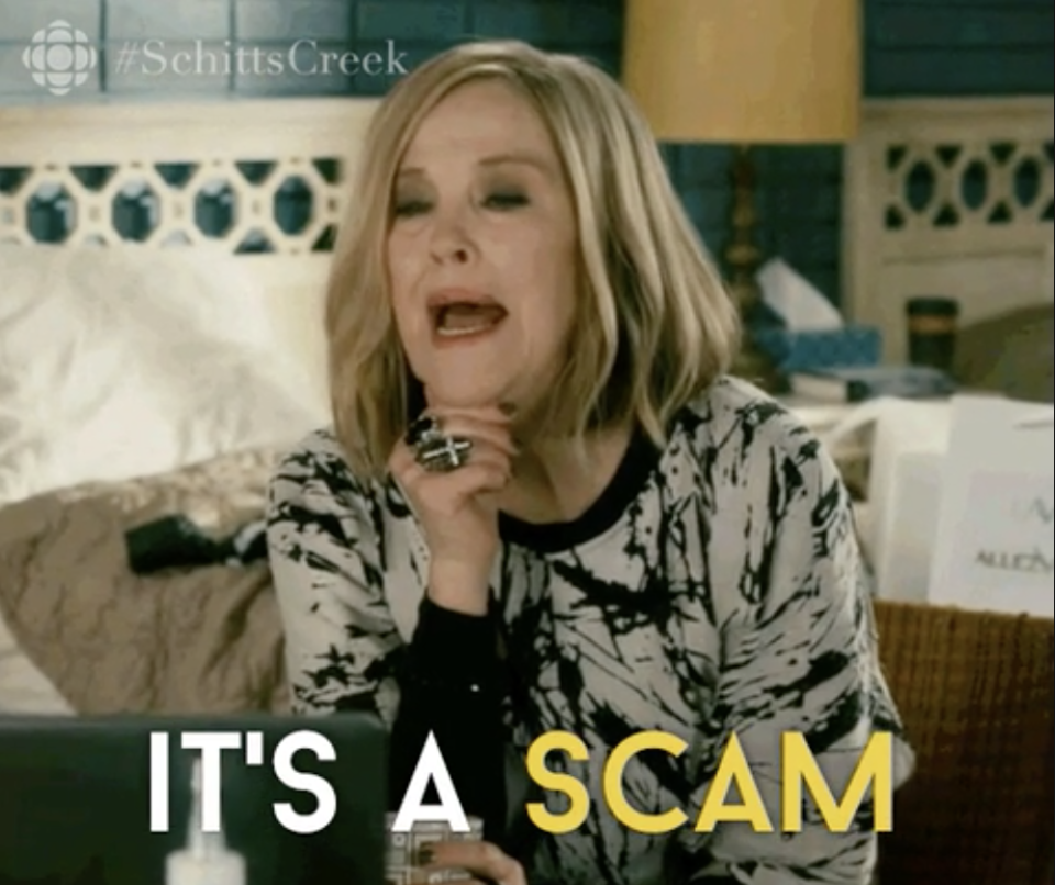 Moira Rose from Schitt's Creek sits at a table, expressive, with text "IT'S A SCAM" overlaying the image