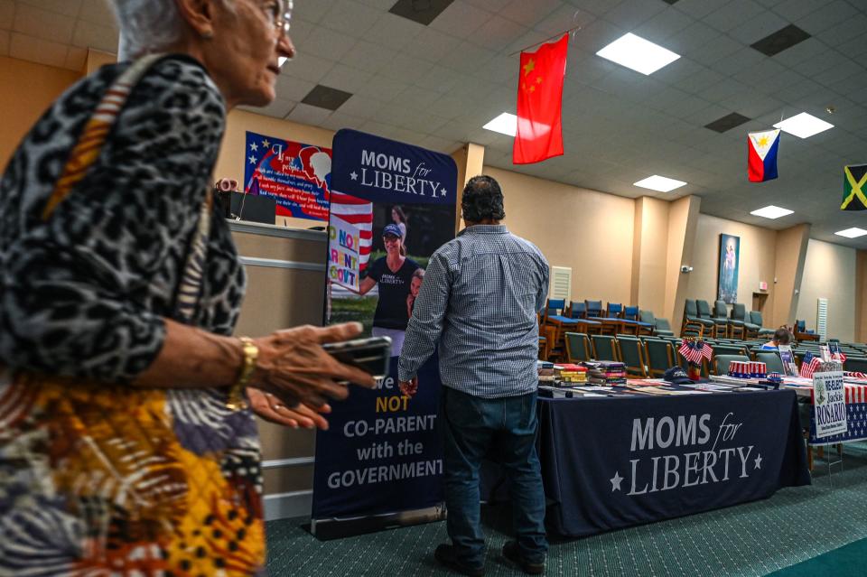 Two people approach a stall of books with a drop cloth saying: Moms for Liberty.