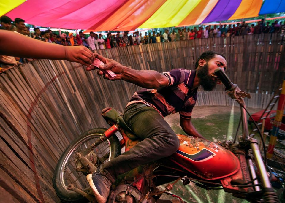 A man riding a motorcycle on the "Well Of Death" grabs a tip offered by an audience member as he rides past, vertically to the ground.