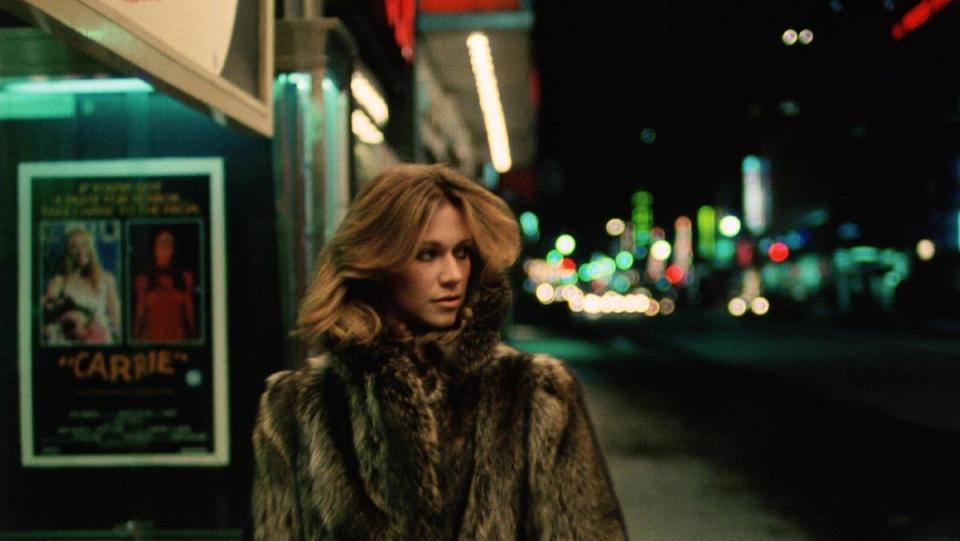 A woman with long hair and a fur coat walks down a city street 