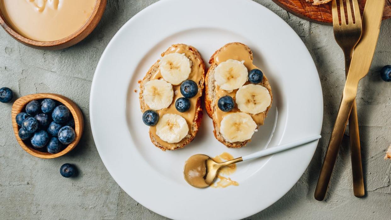 buckwheat healthy bread with peanut butter, banana and blueberry on white plate over concrete background top view flat lay summer breakfast