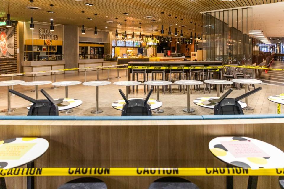 Pictured is an empty food court with upside-down chairs surrounded by a yellow caution tape. 