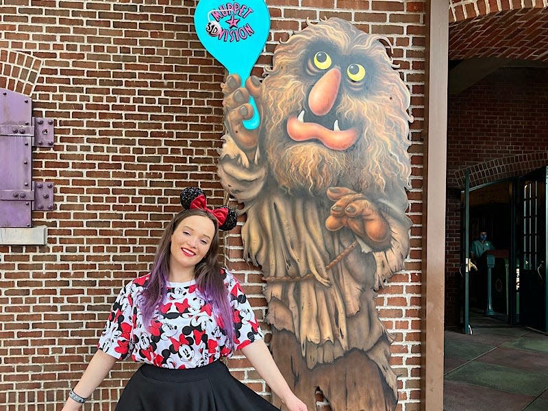 The writer wears a shirt with Minnie Mouse on it, a black skirt, and mouse ears next to a cutout of a Muppet with a sign that says "Muppet 3D Vision"