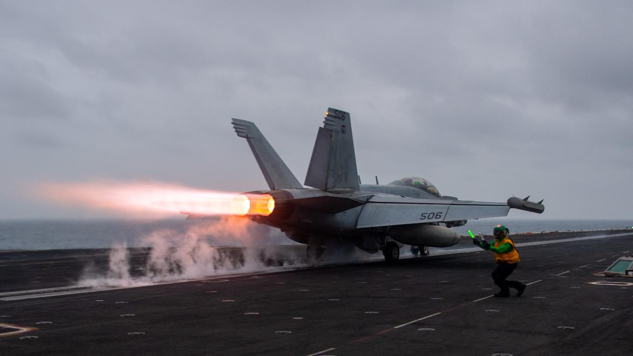 An EA-18G Growler aircraft taking off from the flight deck of a Navy aircraft carrier at sea as a service member in a vest directs it.