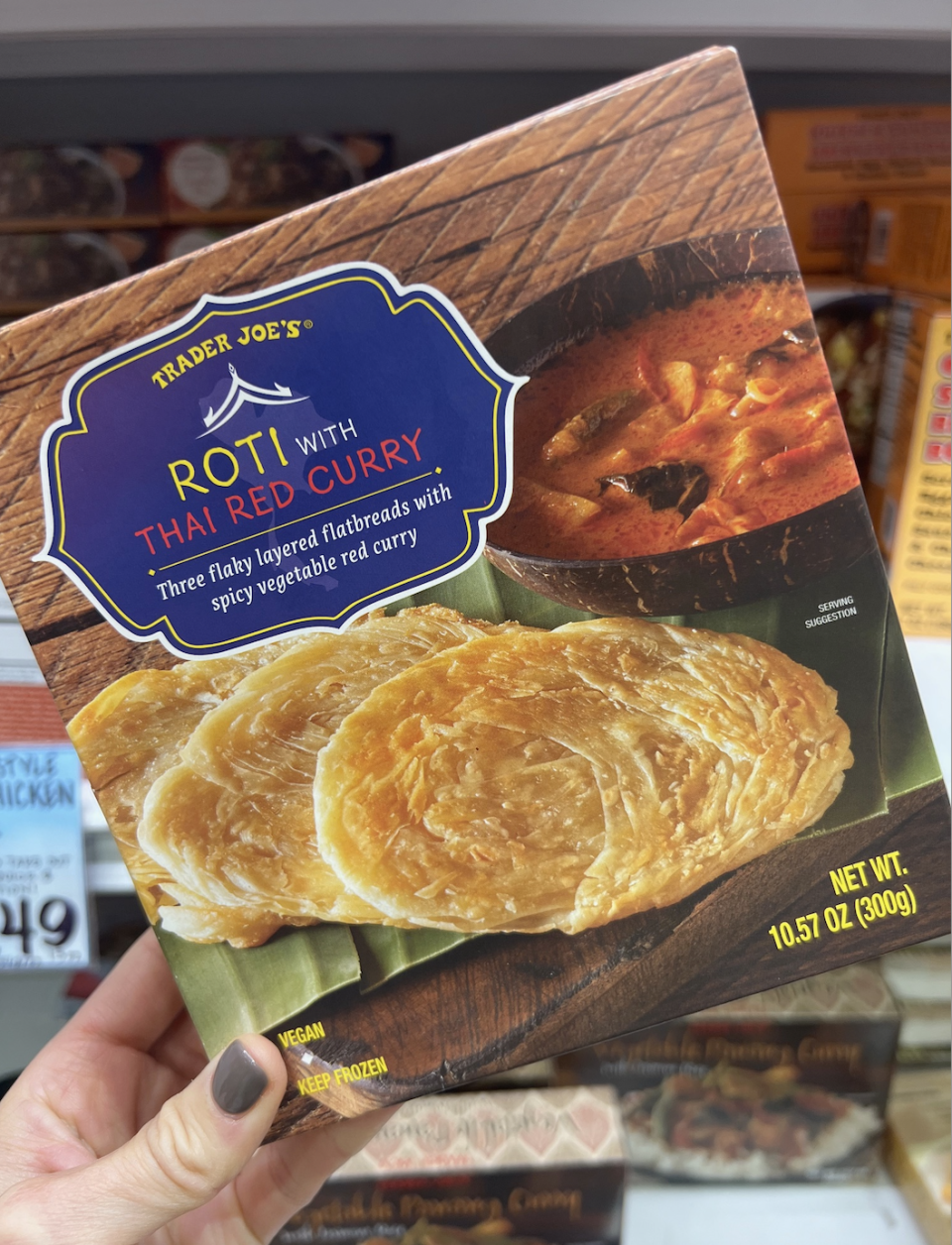 A hand holds a box of Trader Joe's Roti with Thai Red Curry. The package shows three flatbreads and a curry dish, with text highlighting the product's key features