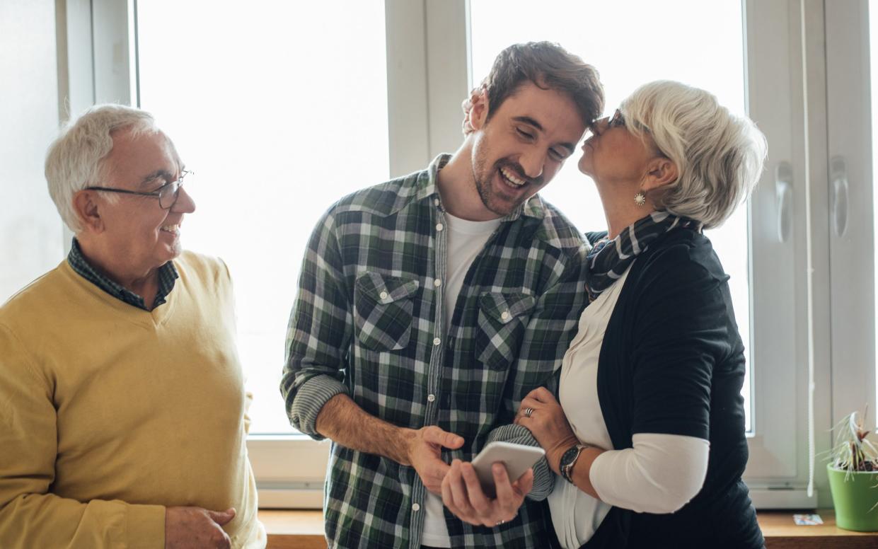 Young man looking at phone, his mother kissing him and father looking at them - Getty