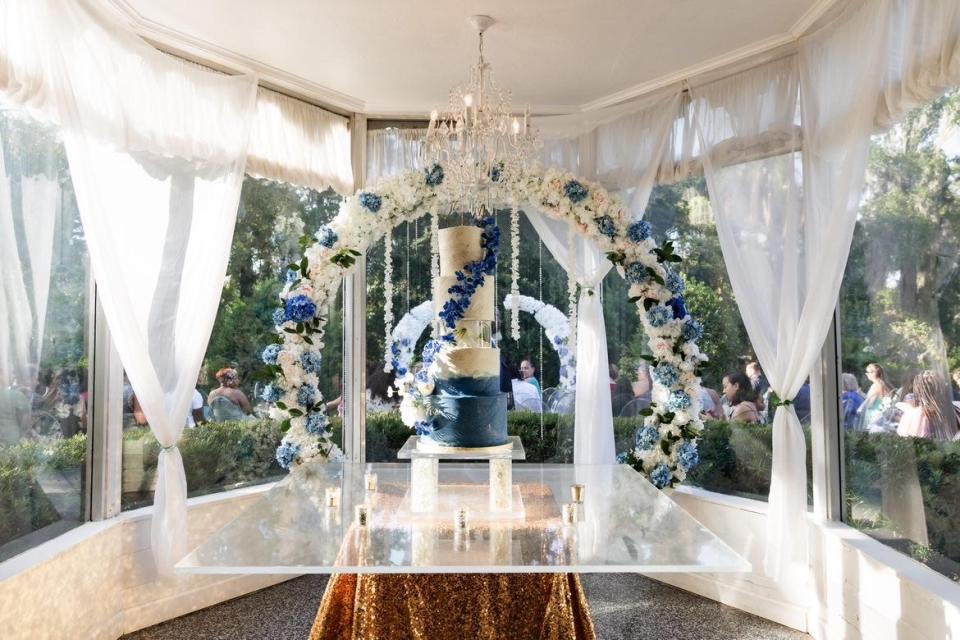 A tall, five-story wedding cake with floral detailing on a clear cake stand in a gazebo with white curtains.