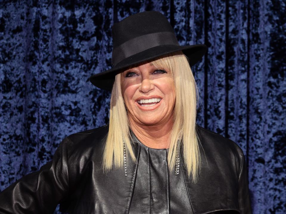 Suzanne Somers smiles at the camera while wearing a black outfit and hat.