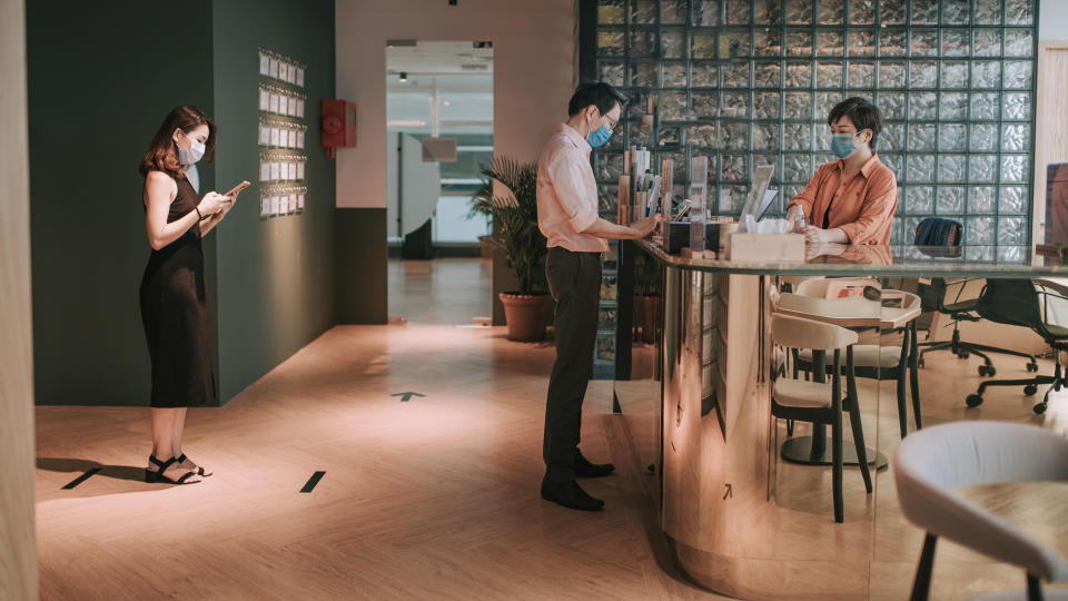 an Asian Chinese receptionist conducting standard operating procedure for illness prevention by measuring visitors colleague coworkers body temperature and record their personal data.