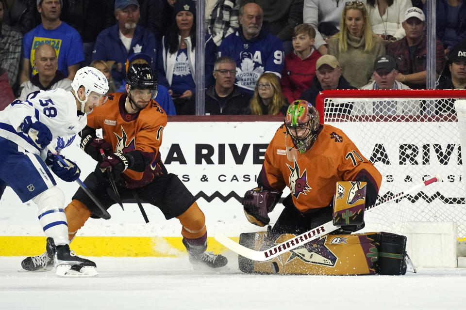 Arizona Coyotes goaltender Karel Vejmelka, right, makes a save on a shot by Toronto Maple Leafs left wing Michael Bunting (58) as Coyotes defenseman Shayne Gostisbehere (14) applies pressure during the first period of an NHL hockey game in Tempe, Ariz., Thursday, Dec. 29, 2022. (AP Photo/Ross D. Franklin)