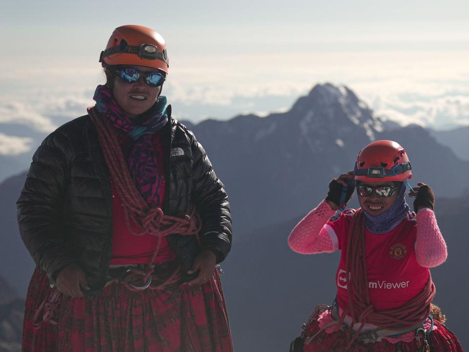 Two Cholita climbers smile at the summit of a mountain.