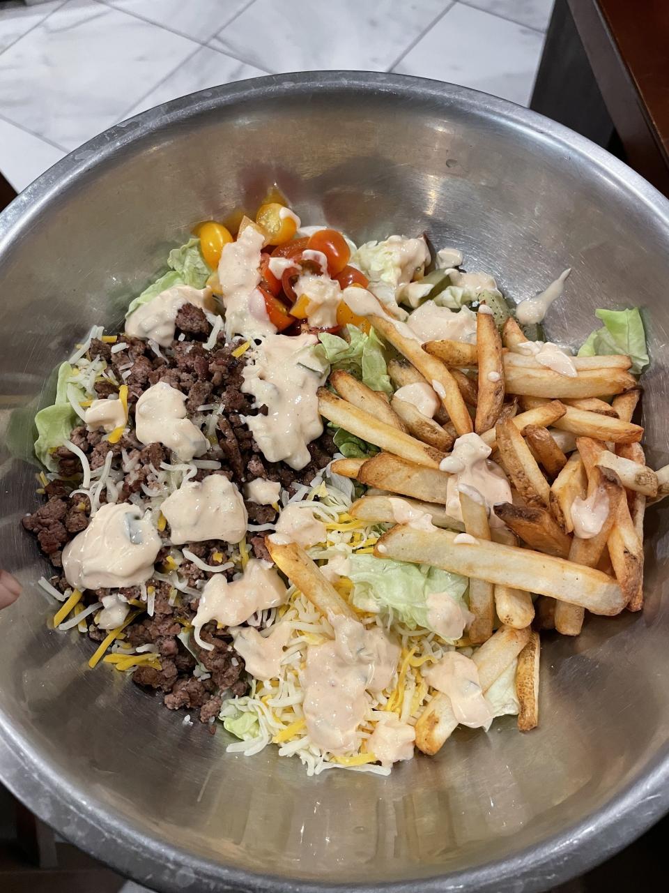 A bowl containing a mixed salad with ground meat, shredded cheese, fries, cherry tomatoes, lettuce, and a creamy dressing