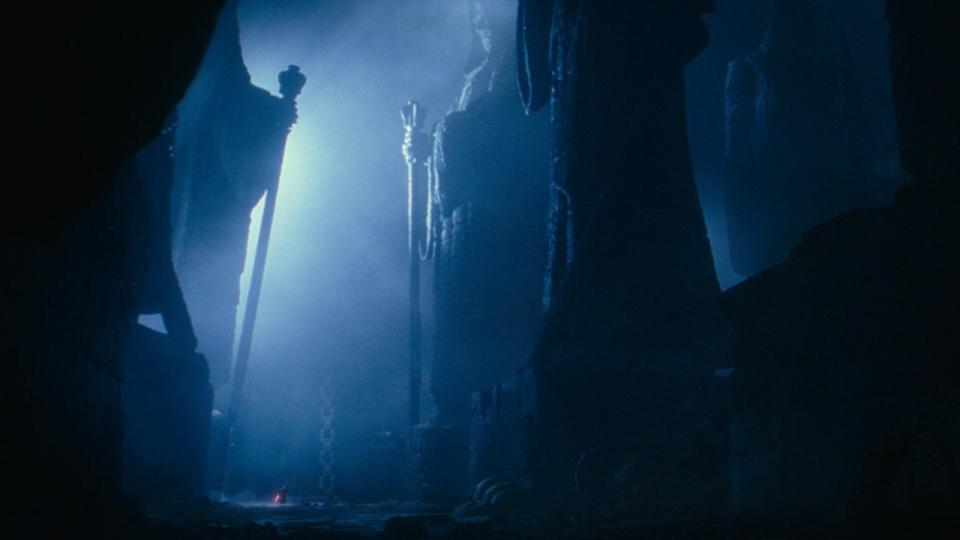 Two giant robed statues holding staffs are at the entrance of the Sith citadel. The scene is in darkness except for the light coming out of the entrance between the two statues.