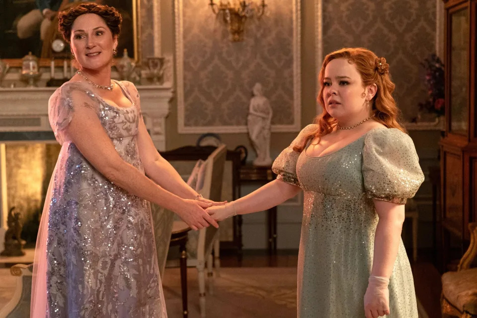 Polly Walker and Nicola Coughlan, in Regency-style dresses, hold hands in a lavishly decorated room.