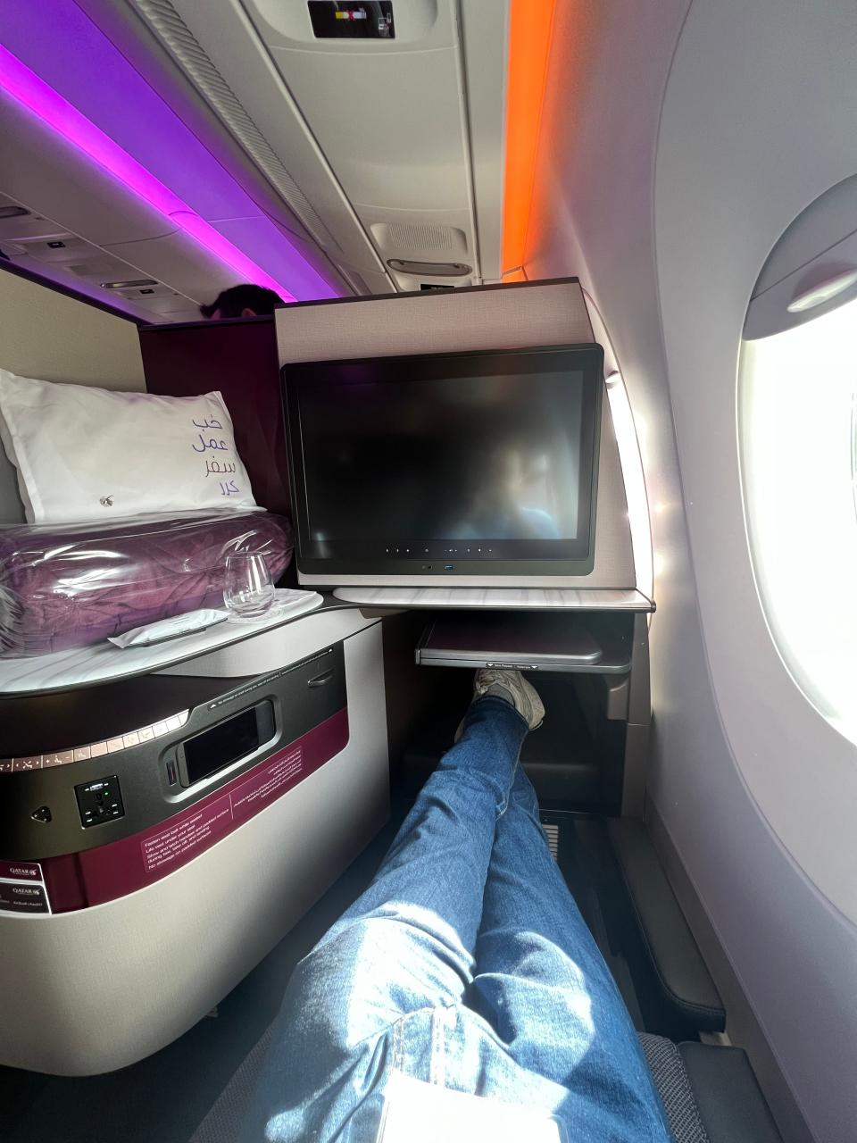 A passenger wearing jeans stretches his legs out on the Qatar A350 business class seat, with the large screen and branded cushions visible