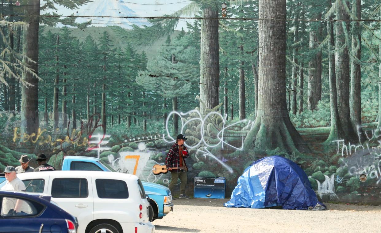 A person with a guitar heads toward a tent pitched in front of the forest mural on the MLK Way facing side of the vacant building planned as a new medical respite facility for Peninsula Community Health Services, in Bremerton on Thursday. The city acknowledges its ordinance that prohibits unauthorized camping is not being enforced, and now the city council is opening a discussion over revisions and other needs that may help address the number of people living unsheltered.