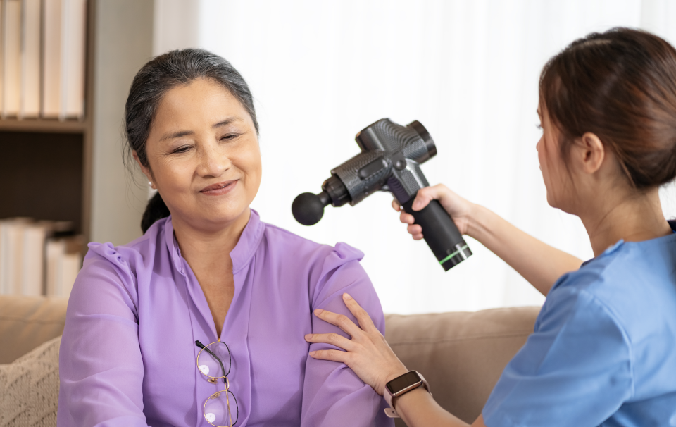 A smiling woman receives a massage from another woman using a handheld massage device on her shoulder