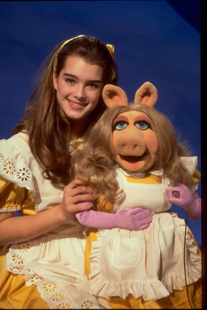 Two characters, Brooke Shields and Miss Piggy from the Muppets, pose in matching yellow dresses with white aprons