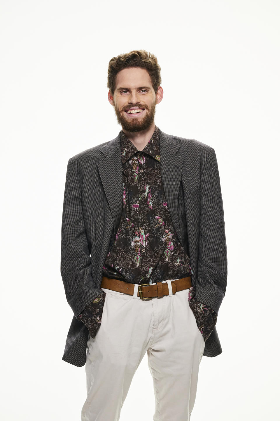 A tall bearded man stands in a dark patterned jacket over a dark shirt with pink patterns, white trousers and tan belt.