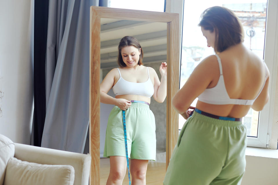 While the allure of a 'goal body' is strong, especially in the age of social media, it's essential to approach body goals with a mindset of health and self-respect. In cozy living room, pretty millennial 35s oversize happy woman in bra and casual shorts holds measure tape check her waist size looking in mirror standing alone at home. Enjoy diet result, weight loss, slimming concept
