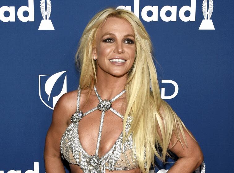 Britney Spears with long hair in a silver, strappy bra top smiling in front of a blue backdrop