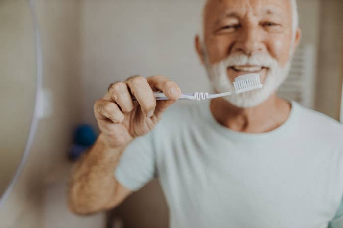 Een oudere man glimlacht terwijl hij een tandenborstel bij zijn mond houdt, wat de mondhygiëne demonstreert