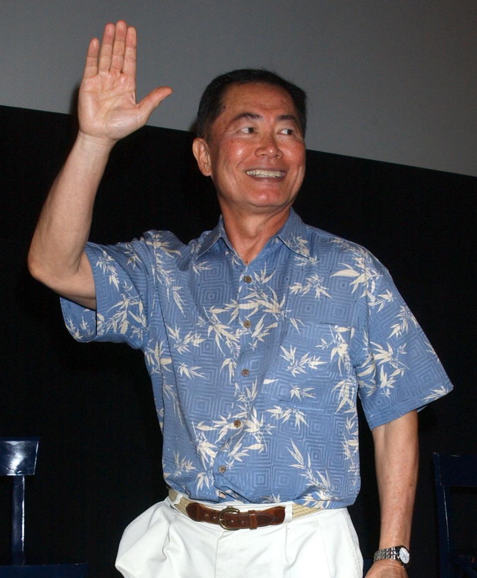 George Takei waves while wearing a patterned short-sleeve shirt, white pants, and brown shoes, holding a water bottle