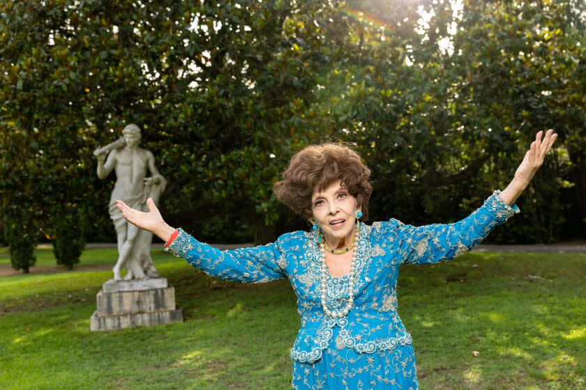 An older woman in a blue dress spreads her arms widely while standing outdoors near a statue
