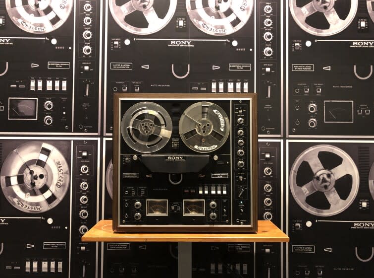 A reel-to-reel tape recorder is seen on a shelf before a wall covered in a wallpaper showing reel-to-reel recorders
