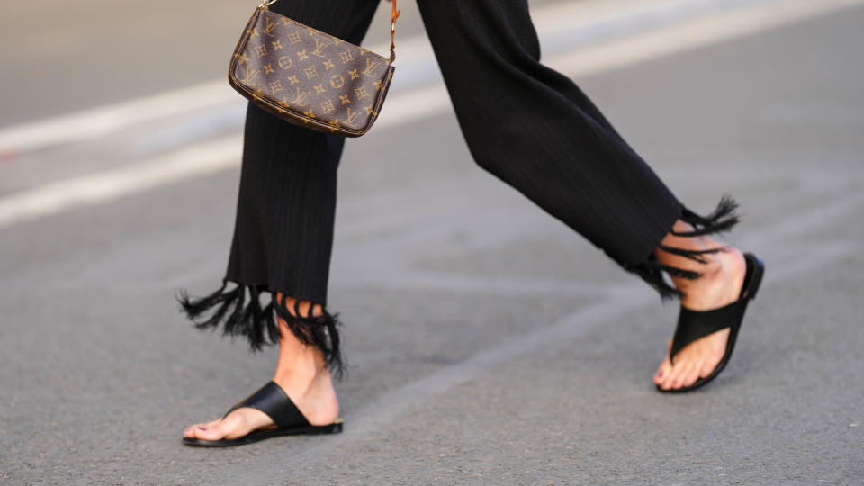 Amanda Derhy wears a brown shiny monogram print pattern in coated canvas shoulder bag from Louis Vuitton, black fringed wide legs pants, black shiny leather flip-flop / shoes, during a street style fashion photo session, on April 17, 2022 in Paris, France.