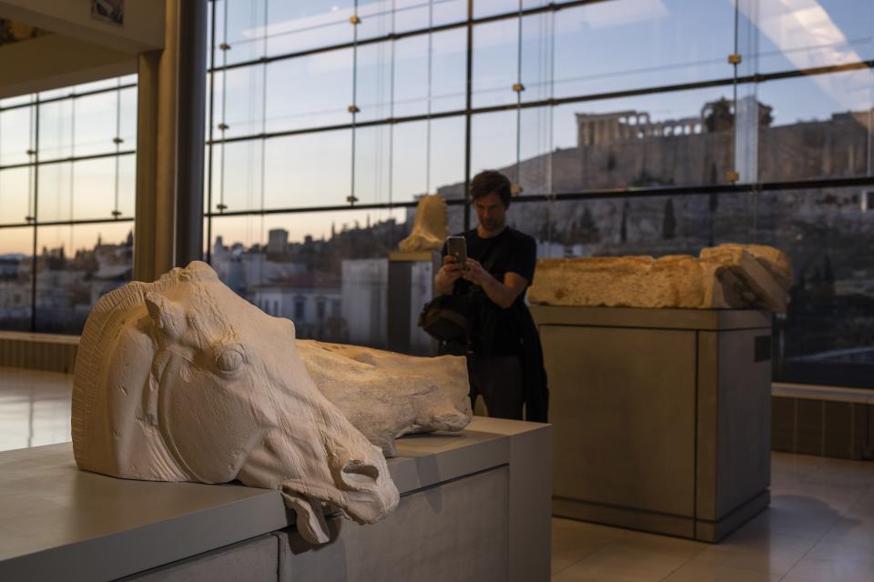 FILE - A man takes a picture of the four-horse chariot of Selene at the Acropolis Museum with the ancient Parthenon temple in background during a ceremony for the repatriation of three sculpture fragments in Athens, on March 24, 2023. Greece's prime minister said Thursday May 11, 2023 his government is exploring a “win-win” solution to one of the world’s most intractable cultural heritage disputes: The fate of the Parthenon Sculptures currently in the British Museum. (AP Photo/Petros Giannakouris, File)
