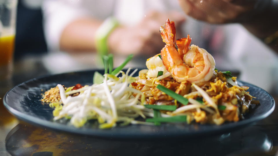 Plate of shrimp pad thai with bean sprouts and garnishes, chef in background