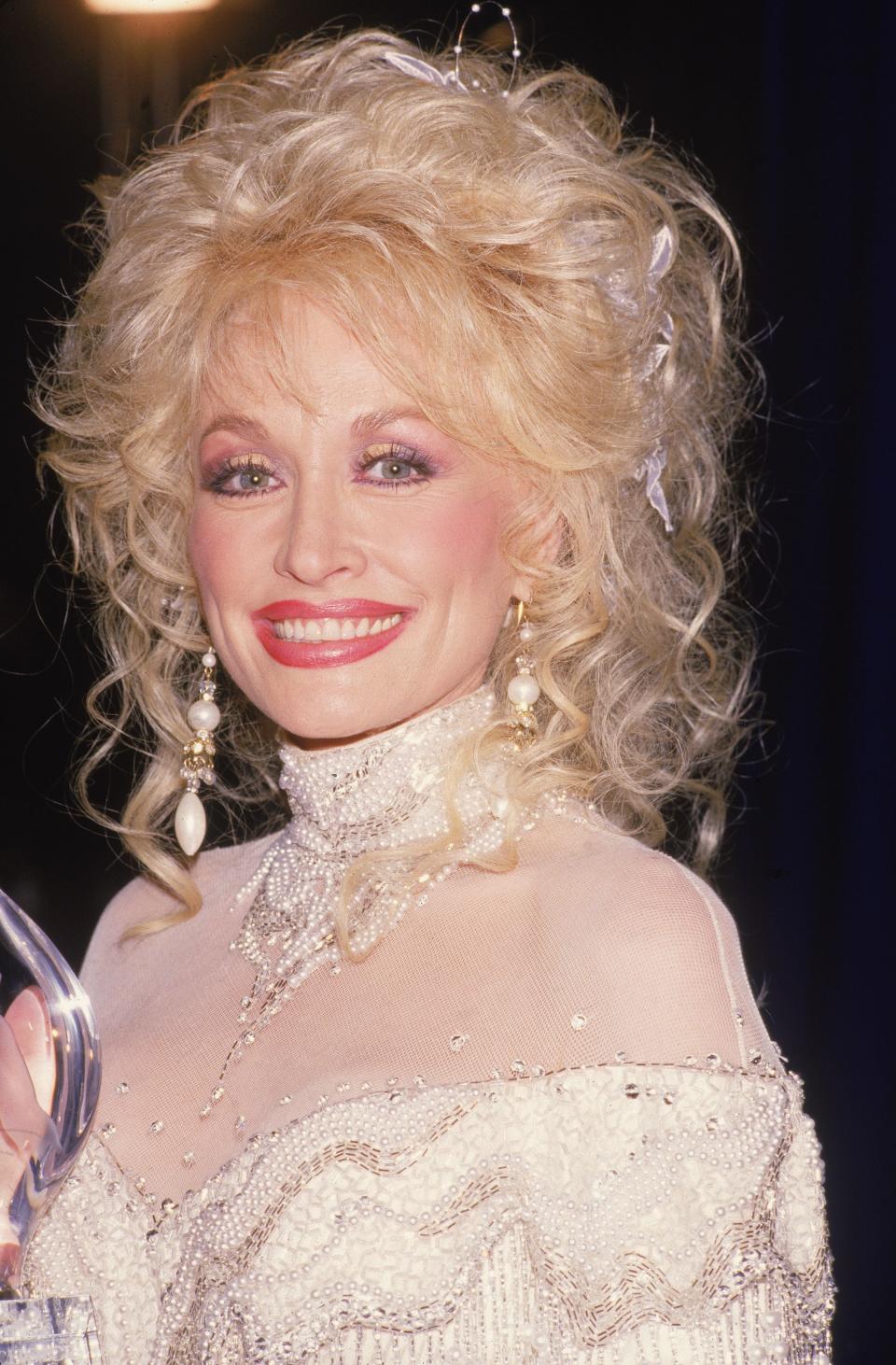 American country music singer and actress Dolly Parton poses with a People's Choice Award, March 13, 1988. She wears a white sequinned dress with transparent shoulders and long earrings. (Photo by Fotos International/Robert Scott/Getty Images)