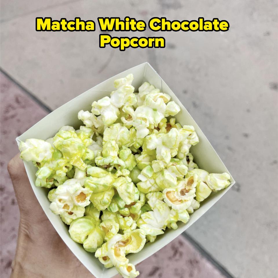 A hand holding a box of popcorn with a green tint, extended over a concrete surface