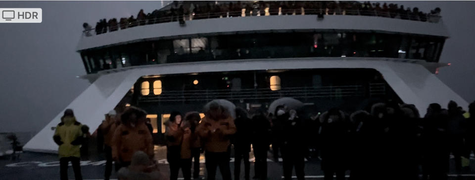 a dark image of a ship with people standing out on the deck and thick gray clouds above.