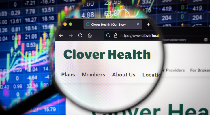 Clover Health (CLOV) company logo on a website with blurry stock market developments in the background, seen on a computer screen through a magnifying glass.