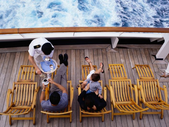 Passengers sit in wooden chairs on the cruise deck while white uniformed staff take care of them