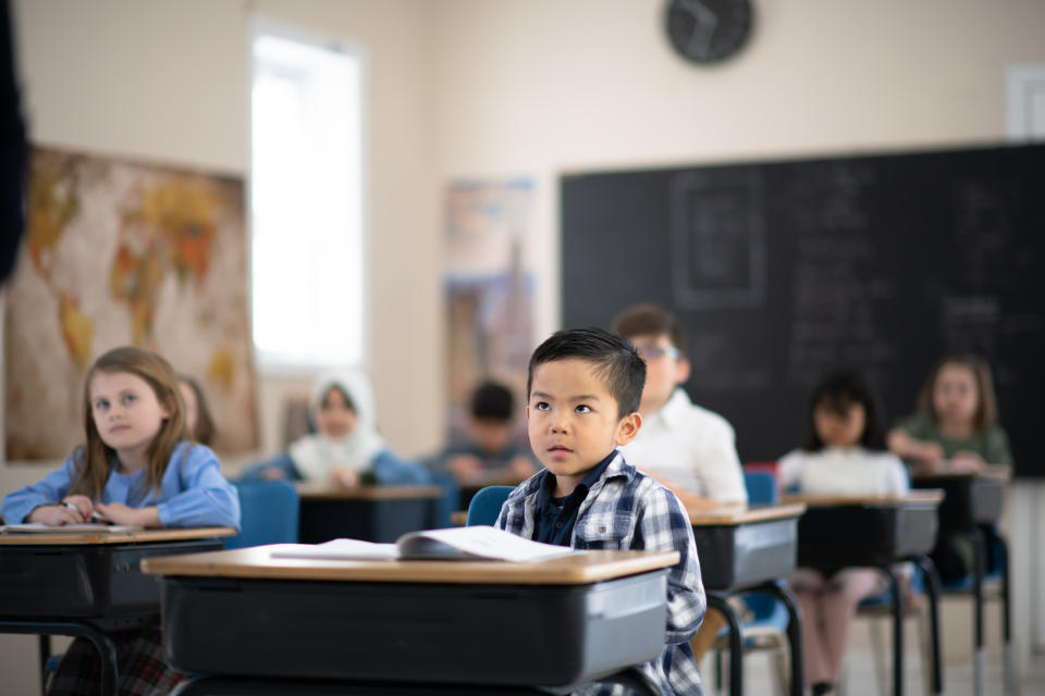 An adorable group of young multi-ethnic children is sitting in class and listening to their teacher speak. All their attention is focused on the front of the room where the teacher is standing.