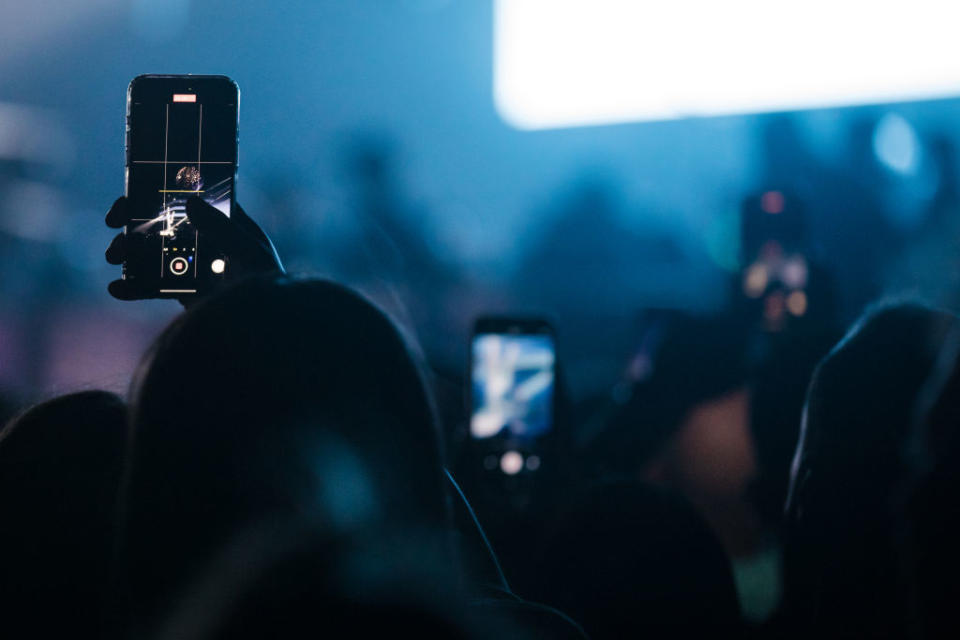 Audience with phones raised at a concert, capturing the event. Focus on one phone's screen showing the stage
