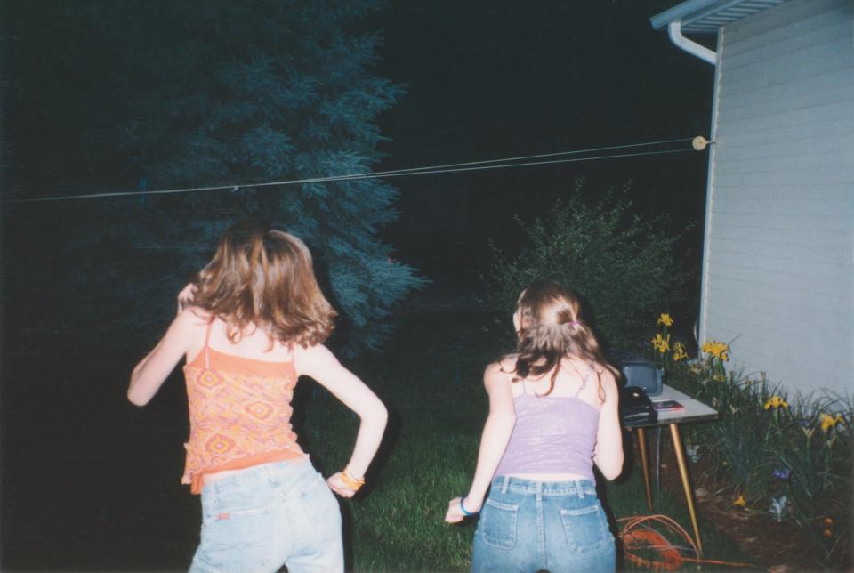 Two people, seen from behind, are dancing outdoors at night near a house. They are casually dressed in jeans and tops