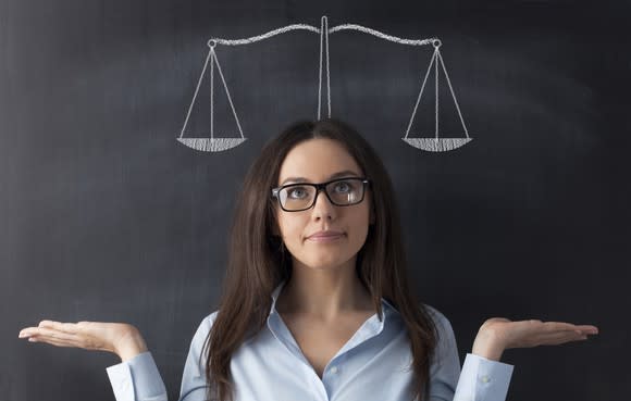 A woman stands with her back against a chalkboard, holding her palms up and looking at a drawing of a balance scale above her head.