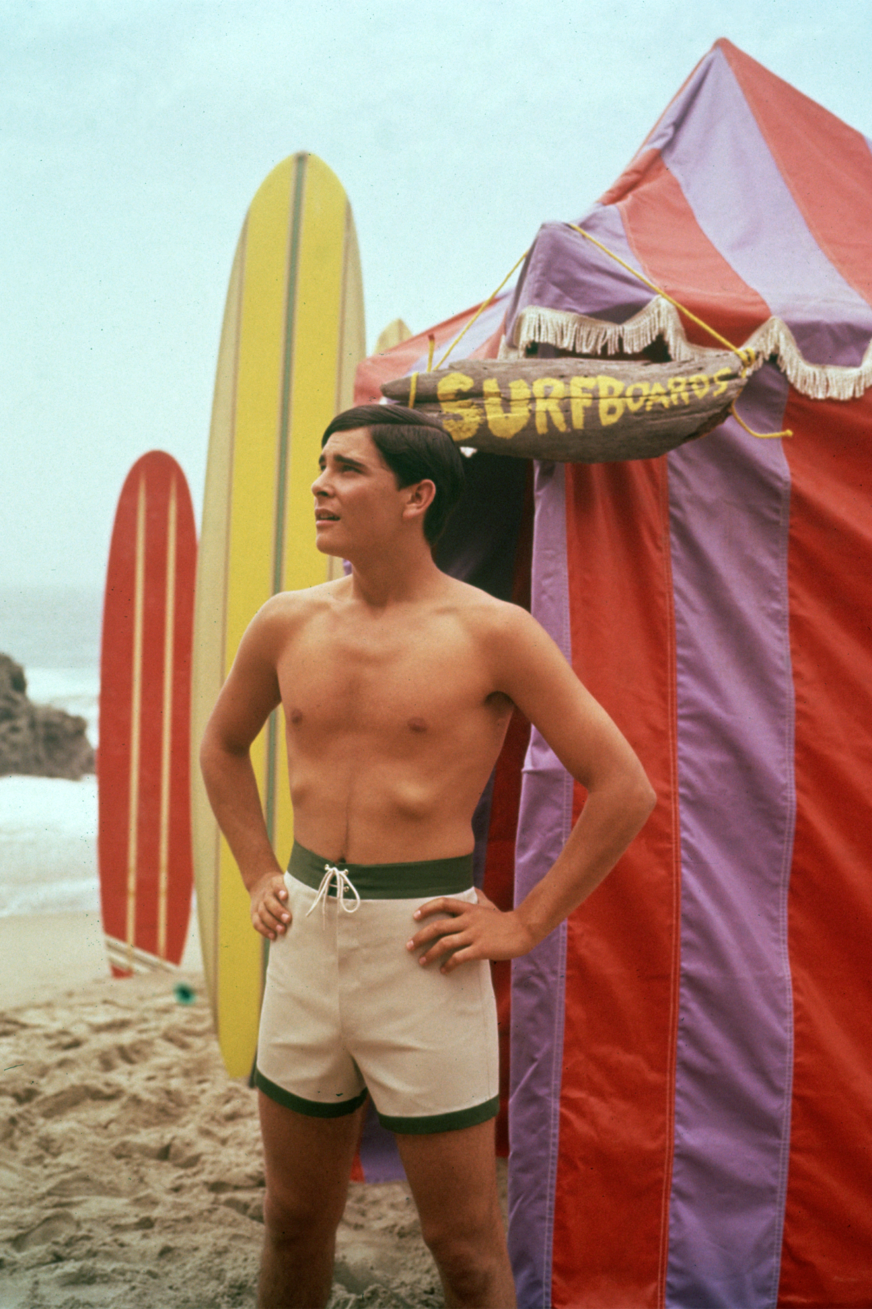 A man wearing swimming trunks standing outside a beach hut with a sign that says, 'Surfboards', with two surfboards sticking out of the sand