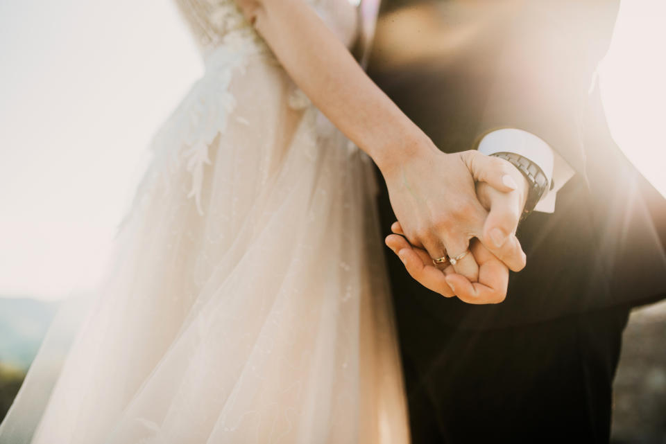 A bride and groom hold hands tightly, showcasing their wedding rings. The bride's lacy dress and the groom's suit sleeve with cufflinks are visible