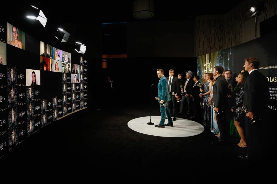 A group of actors stand in front of screens of reporters watching remotely